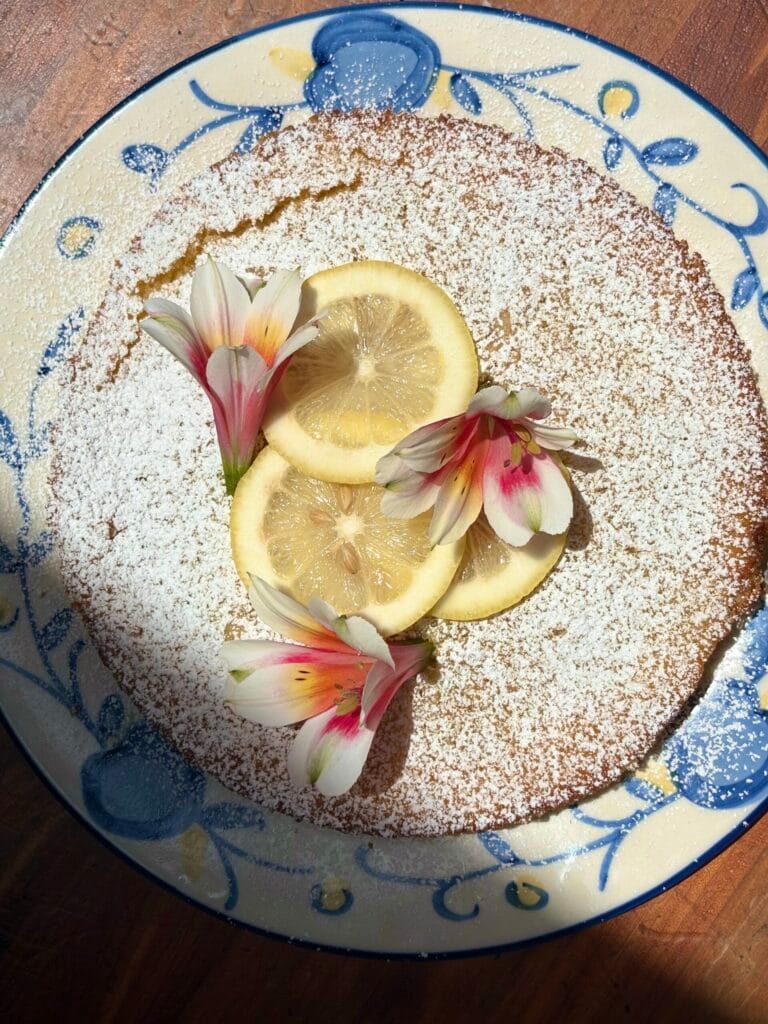 This image shows a Lemon & Olive Oil cake decorated with lemon wedges and flowers.