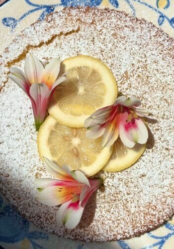 This image shows a Lemon & Olive Oil cake decorated with lemon wedges and flowers.